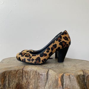 May include: A pair of high-heeled pumps featuring a textured leopard print pattern in shades of brown, tan, and black. The shoes have a rounded toe, a black interior lining, and a solid black block heel that provides a stable base for the footwear.