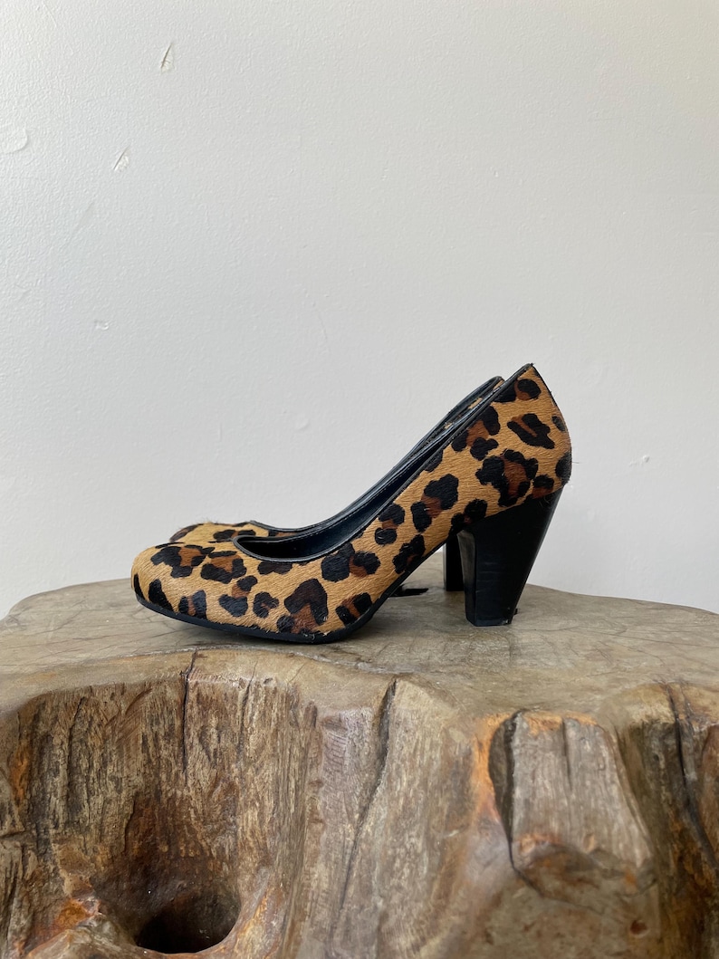 May include: A pair of high-heeled pumps featuring a textured leopard print pattern in shades of brown, tan, and black. The shoes have a rounded toe, a black interior lining, and a solid black block heel that provides a stable base for the footwear.