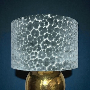 May include: A teal lampshade with a leopard print pattern sits atop a gold-colored lamp base. The lampshade is cylindrical and made of a soft, textured material. The lamp base has a hammered metal finish.