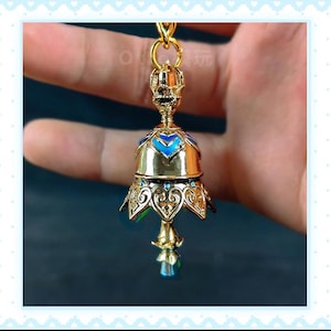 Op de afbeelding: Een goudkleurige, klokvormige hanger met blauwe emaille accenten en een heldere, facetgeslepen kristal aan de onderkant. De hanger heeft ingewikkelde filigraan details en is opgehangen aan een gouden ketting.