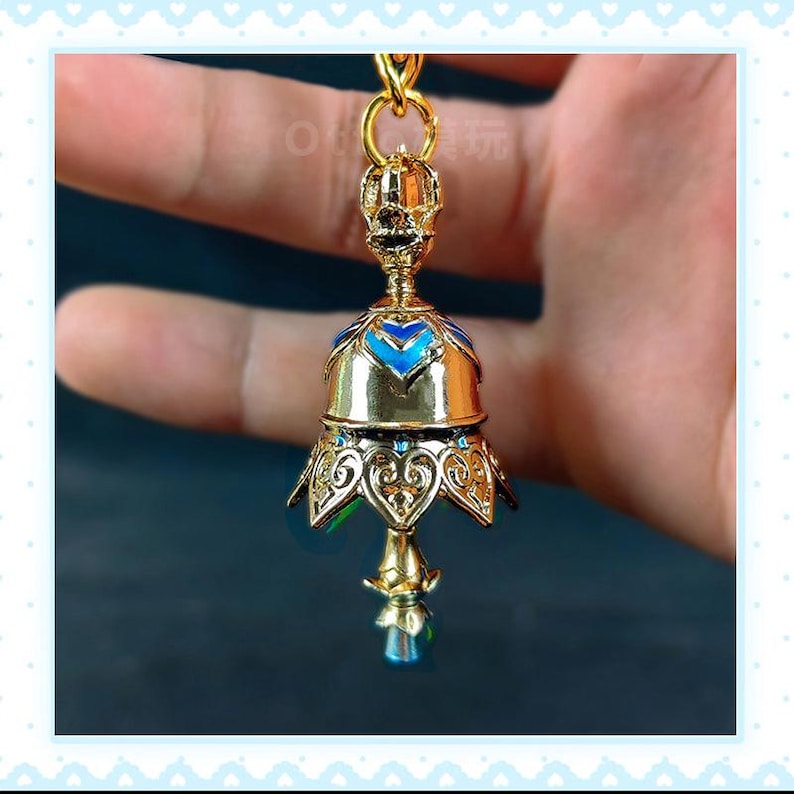 Op de afbeelding: Een goudkleurige, klokvormige hanger met blauwe emaille accenten en een heldere, facetgeslepen kristal aan de onderkant. De hanger heeft ingewikkelde filigraan details en is opgehangen aan een gouden ketting.