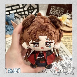May include: A plush doll with brown fur, embroidered facial features, and a red and black outfit. The doll has long braids and is holding a small red cape. The text "粉" and "【孙策】" is visible.