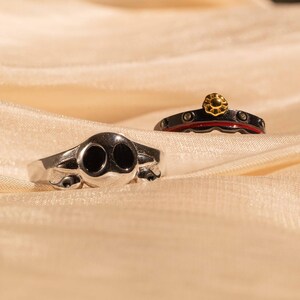 May include: Two silver rings on a beige fabric background. One ring has a skull design with black eyes. The other ring has a red and black band with a gold flower design.