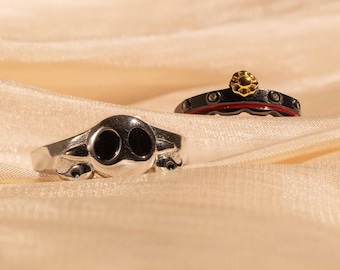 Könnte beinhalten: Zwei silberne Ringe auf einem beigefarbenen Stoffhintergrund. Ein Ring hat ein Totenkopfdesign mit schwarzen Augen. Der andere Ring hat ein rot-schwarzes Band mit einem goldenen Blumendesign.