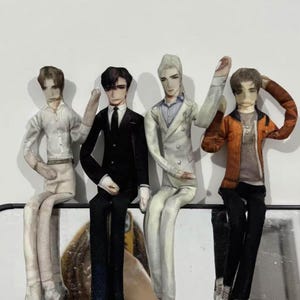 May include: Four fabric dolls of men in different outfits. One doll is in a white shirt and beige trousers, another in a black suit and tie, a third in a white suit, and the last in an orange jacket and black trousers. They are seated.