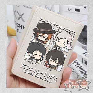 Puede incluir: Cartera beige con ilustraciones de personajes de dibujos animados y texto japon&eacute;s, incluyendo "Bungo Stray Dogs". La cartera tiene aproximadamente 0,8 cm de grosor. Tambi&eacute;n se ve el texto "GUNGO STRAY DOGS".