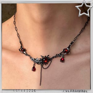 May include: A dark silver necklace with red gemstone accents. The necklace features a central spiky design with dangling teardrop-shaped gems and chain details. The necklace is worn around the neck.