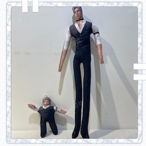 May include: Two fabric dolls, one large and one small, both wearing black waistcoats, white shirts, and black trousers. The larger doll has a black choker. The dolls have light skin and styled hair. The smaller doll has its arms outstretched.