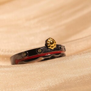 May include: A black and red ring with a gold flower design and small clear stones. The ring is on a light brown fabric background.