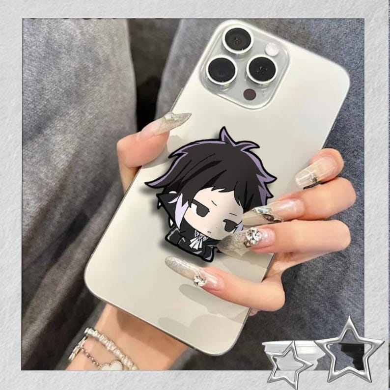 May include: A white smartphone with a cartoon character phone grip. The grip features a chibi-style anime character with black hair and a black outfit. The phone is held in a hand with long, decorated nails.