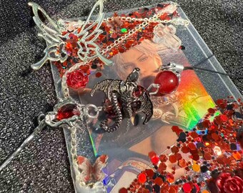 Puede incluir: Una funda rectangular transparente con temática de fantasía, con un retrato, purpurina roja y abalorios metálicos decorativos. Los abalorios incluyen una mariposa, un dragón y una rosa. Un colgante de orbe rojo cuelga de un cordón negro.