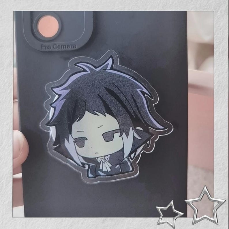 May include: A clear acrylic phone grip featuring a cartoon character with black hair and a white collar. The character has a neutral expression and is set against a black phone case. The phone case has a camera bump that says "Pro Camera".