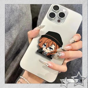 May include: A white smartphone with a cartoon character sticker featuring a character with brown hair and a black hat. The phone has a triple-lens camera and is held by a person with long, decorated nails.