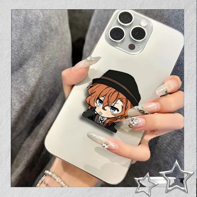 May include: A white smartphone with a cartoon character sticker featuring a character with brown hair and a black hat. The phone has a triple-lens camera and is held by a person with long, decorated nails.