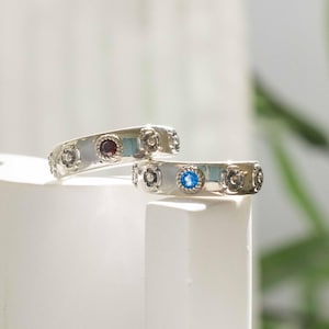May include: Two silver rings with floral designs and colored gemstones. One ring features a red gemstone, the other a blue gemstone. The rings are displayed on a white surface, with a soft background.