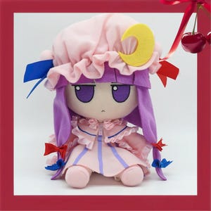 Op de afbeelding: Een pluche pop met een roze jurk en hoed, paars haar en een gele halve maan. De pop heeft grote paarse ogen en is versierd met blauwe en rode strikken. De pop is een zacht, knuffelig speelgoed.