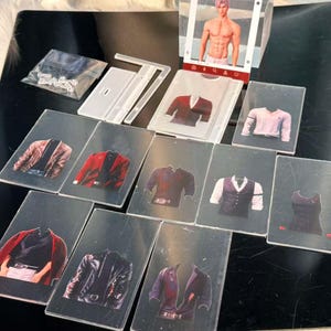 May include: A collection of fashion-themed cards featuring various jacket and shirt designs. The cards showcase a range of colors, including red, black, and brown, with detailed illustrations of clothing styles. A display stand with a male figure is also present.