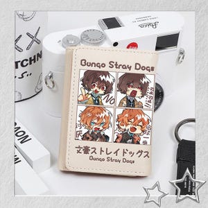 Puede incluir: Una cartera beige con una ilustraci&oacute;n de estilo manga de personajes de "Bungo Stray Dogs". La cartera tiene el t&iacute;tulo en ingl&eacute;s y japon&eacute;s. Una c&aacute;mara blanca y otros accesorios est&aacute;n en el fondo.