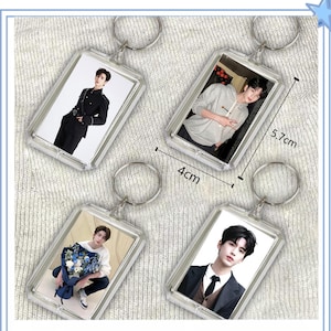 May include: Four rectangular clear acrylic keychains featuring photographic portraits of a person. Each keychain includes a metal ring. The dimensions are clearly marked as 4 cm by 5.7 cm. The images show a person in various outfits, including a black uniform, a grey hoodie, a white sweater with a blue floral bouquet, and a dark suit with a tie.