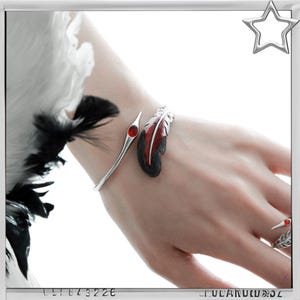 Puede incluir: Un brazalete de pluma plateada con un dise&ntilde;o de pluma roja y negra y un anillo a juego con una piedra roja. El brazalete se envuelve alrededor de la mu&ntilde;eca y el anillo est&aacute; en el dedo anular. La joyer&iacute;a est&aacute; sobre un fondo blanco.