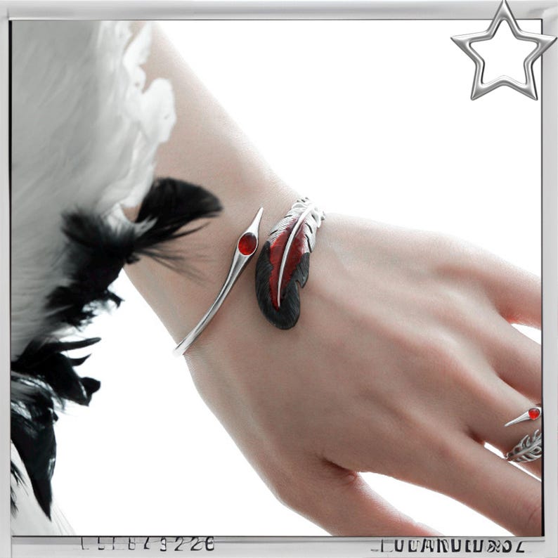 Puede incluir: Un brazalete de pluma plateada con un dise&ntilde;o de pluma roja y negra y un anillo a juego con una piedra roja. El brazalete se envuelve alrededor de la mu&ntilde;eca y el anillo est&aacute; en el dedo anular. La joyer&iacute;a est&aacute; sobre un fondo blanco.