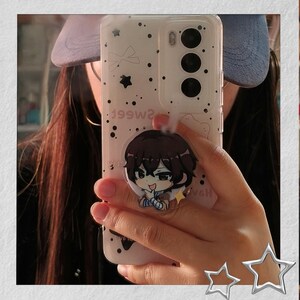 May include: Clear phone case with a cartoon character pop socket. The case has a white background with black dots and stars. The pop socket features a brown-haired anime character with a blue and white outfit.