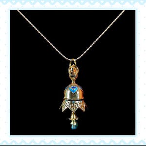 Op de afbeelding: Een goudkleurige, klokvormige hangerketting met blauwe emaille accenten en een kleine blauwe edelsteen aan de onderkant. De hanger hangt aan een fijne zilveren ketting tegen een zwarte achtergrond.