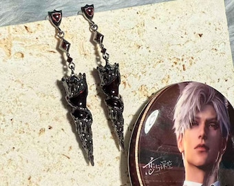 May include: A pair of long, dark gray earrings with red accents. Each earring features a shield-shaped top, a series of diamond-shaped charms, and a stylized, twisted bottom. A round button with a male anime character is also visible.