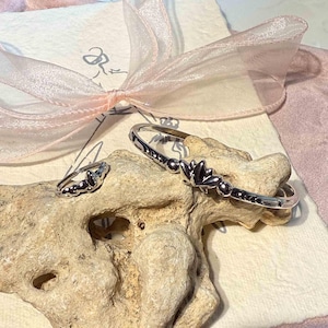 Könnte beinhalten: Ein silbernes Armband- und Ringset mit floralem Design. Das Armband ist im Bangle-Stil gehalten, und der Ring hat einen passenden floralen Akzent. Der Schmuck wird auf einem Stück Treibholz mit rosa Schleife und cremefarbenem Hintergrund präsentiert.