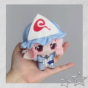 Pode incluir: Uma boneca de pelúcia com uma personagem de cabelo rosa, olhos vermelhos e uma roupa azul. A boneca usa um chapéu azul com aba branca e um desenho de redemoinho vermelho. A boneca é segurada em uma mão.