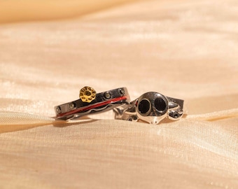 Könnte beinhalten: Zwei silberne Ringe mit schwarzem und rotem Email. Ein Ring hat ein kleines goldenes Sonnenmotiv, der andere einen Totenkopf.