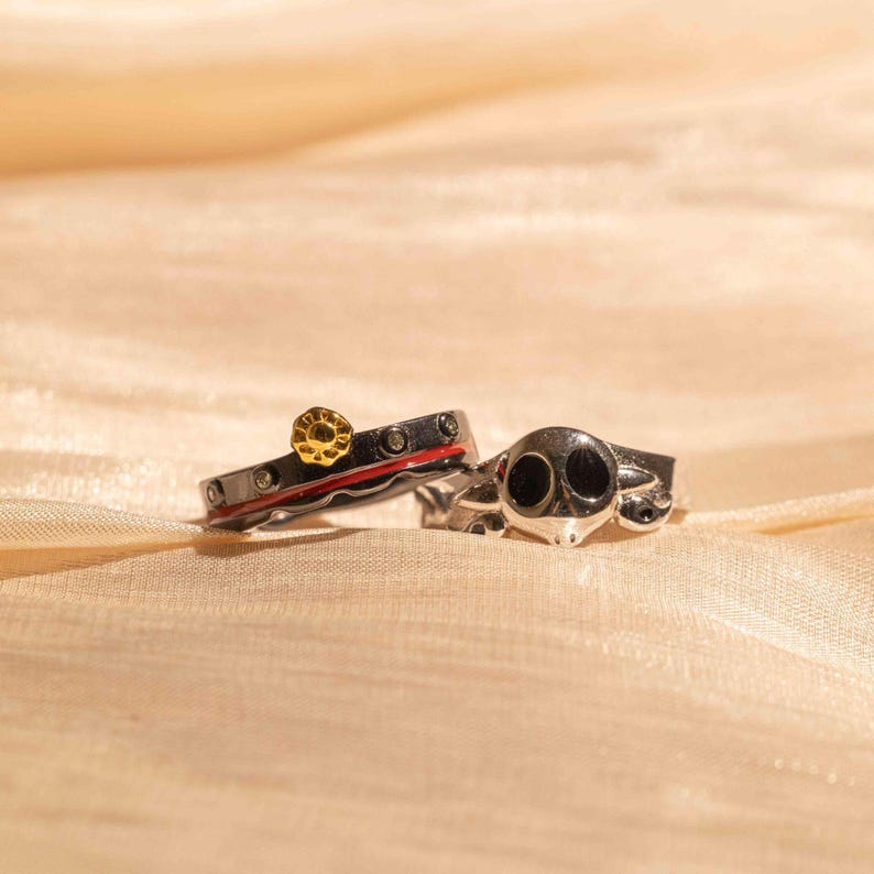 May include: Two silver rings with black and red enamel. One ring features a small gold sun design and the other ring features a skull design.