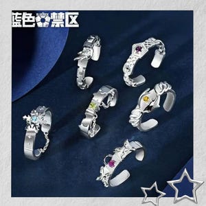 Può includere: Una collezione di anelli argentati con vari design, tra cui draghi e forme geometriche, ognuno ornato con pietre preziose colorate. Gli anelli sono esposti su una superficie blu scuro, con il testo "Blue Lock" nell'angolo in alto a sinistra.