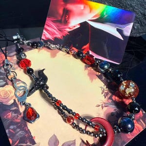 May include: A gothic-style beaded necklace. It features black, red, and silver beads, a black raven charm, and a red crescent moon pendant. Additional charms include a handcuff and a red gem.