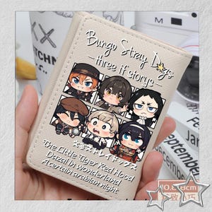 Puede incluir: Una cartera beige con ilustraciones de dibujos animados de personajes de "Bungo Stray Dogs". La cartera tiene el t&iacute;tulo de la serie y frases como "three if stories" y "The Little Tiger Red Hood" impresas. La cartera mide aproximadamente 10,8 cm.