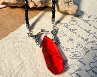 May include: A red crystal pendant necklace with a black braided leather cord. The pendant is a faceted, elongated crystal shape, held by a silver-toned metal setting. The necklace rests on a paper with handwritten text.