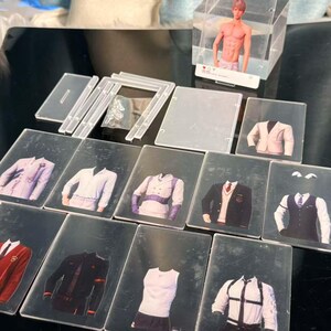 May include: A collection of clear acrylic display pieces and cards featuring various outfits. The cards showcase different jacket and shirt combinations, with a small figure in a clear box in the background. The outfits include blazers, vests, and casual wear.