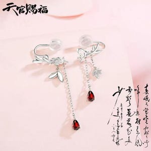 May include: A pair of silver-toned earrings with floral and leaf designs. One earring features a long chain with a teardrop-shaped, red gemstone. The other earring has a shorter chain with a red gemstone and a maple leaf charm.