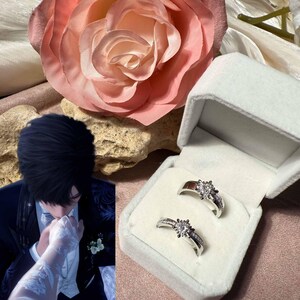 Puede incluir: Dos anillos de plata, cada uno con una piedra central grande y transparente y piedras de acento más pequeñas, se muestran en una caja blanca. Una gran rosa rosa y una persona con ropa formal están en el fondo.