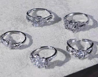May include: Five silver rings with intricate floral and geometric designs. Each ring features sparkling clear gemstones, creating a delicate and elegant appearance. The rings are displayed on a light-colored surface.