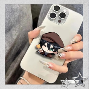 May include: A white smartphone with a cartoon character sticker. The sticker features a character with black hair, green eyes, and a brown hat and coat. A small yellow star is next to the character. The phone is held in a hand with long, decorated nails.
