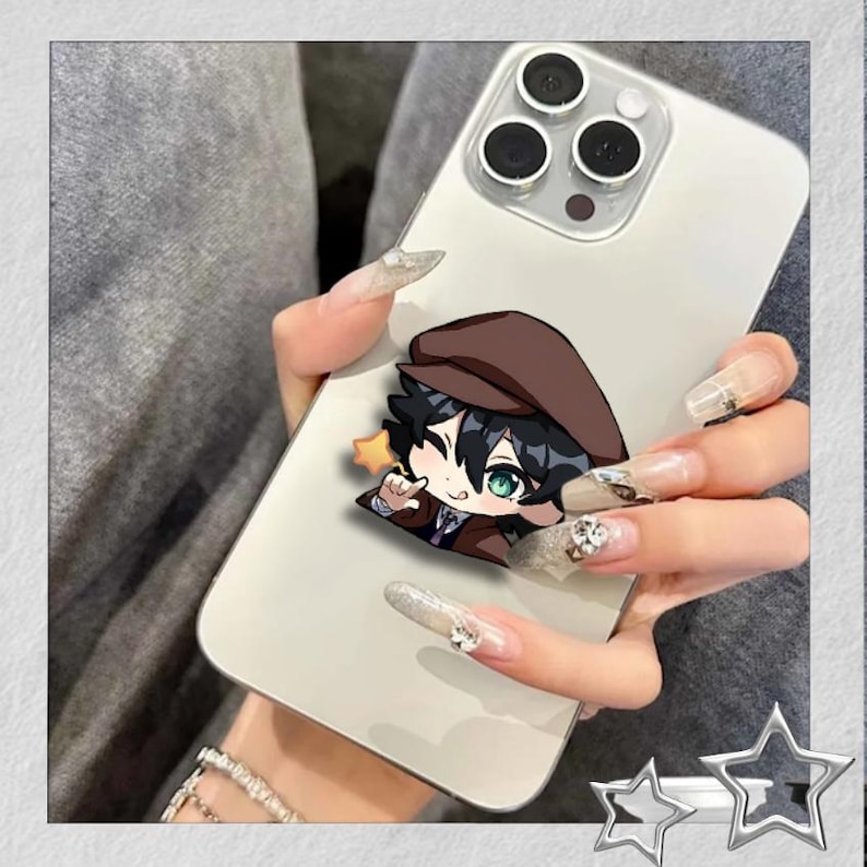 May include: A white smartphone with a cartoon character sticker. The sticker features a character with black hair, green eyes, and a brown hat and coat. A small yellow star is next to the character. The phone is held in a hand with long, decorated nails.