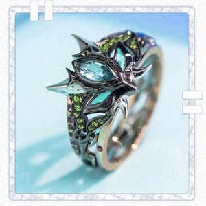 Könnte beinhalten: Ein verzierter Ring mit einem zentralen hellblauen Edelstein, umgeben von kleineren grünen und blauen Steinen. Der Ring hat ein silbernes und goldenes Band mit aufwendigen Details, wodurch ein einzigartiges und elegantes Design entsteht.