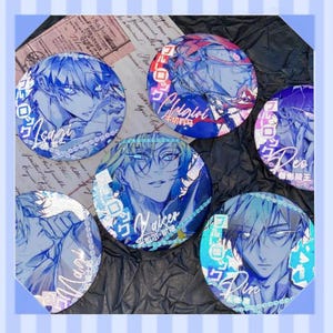 Op de afbeelding: Een verzameling van zes ronde, blauw-witte knopen in anime-stijl. Elke knoop heeft een andere karakterillustratie met Japanse tekst en namen. De knopen zijn gerangschikt op een zwarte, getextureerde ondergrond.