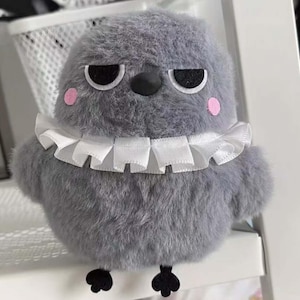 Puede incluir: Un juguete de peluche gris con forma de pájaro, con un cuello blanco con volantes, ojos negros y un pico negro. El pájaro tiene mejillas rosas y pequeños pies negros. El juguete está diseñado para parecerse a un pájaro.