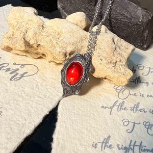 May include: A silver pendant necklace with a bright red gemstone. The pendant has a detailed, textured frame. The necklace is presented on a textured, off-white surface with a rock in the background.