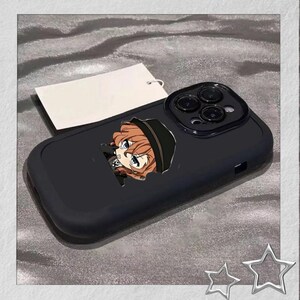 May include: Black phone case featuring a cartoon character with orange hair and a black hat. The case has a matte finish and a camera cutout. A blank white tag is attached to the case.