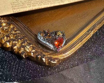 May include: A silver-toned ring with an ornate design, featuring a large, heart-shaped orange gemstone as the focal point. The ring is set on a gold-colored surface, showcasing its intricate details and vibrant colors. The ring's design includes smaller gemstones.