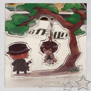 May include: A clear acrylic charm depicting two anime-style characters beneath a tree. One character is suspended from the tree, the other stands nearby. The tree has green foliage and a brown trunk. The charm has a silver keychain.