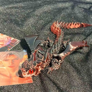 Op de afbeelding: Een gedetailleerd, 3D-geprint drakenbeeldje in rood en zwart. De draak heeft grote vleugels, een lange staart en rode ogen. Het beeldje ligt op een getextureerd oppervlak, met twee kaarten met illustraties in anime-stijl.
